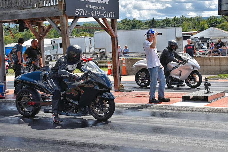 Two motorcyclists lined up for a drag race on a track, one riding a black motorcycle and the other in a white suit on a silver bike, with spectators and a timer in the background.