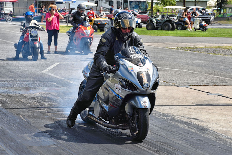 A motorcyclist in leather gear performing a burnout on a racing track, with smoke coming from the rear tire. Other motorcycles and spectators are visible in the background.