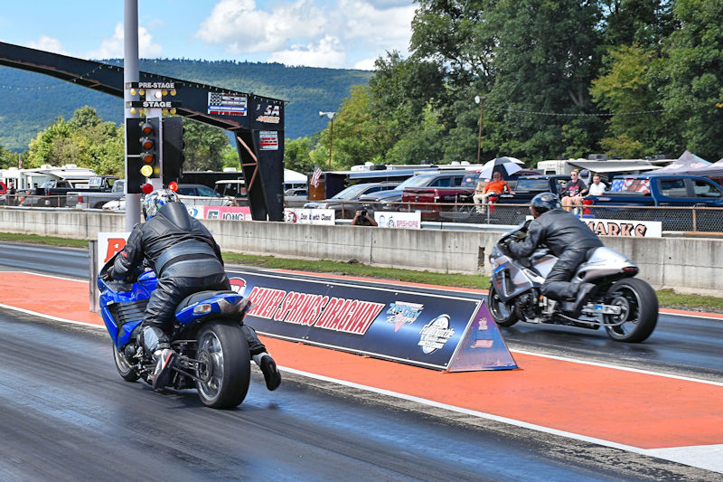 Two motorcycle racers are competing on a drag strip, with one rider on a blue bike and the other on a silver bike, approaching the starting line. The background features trees and parked vehicles under a partly cloudy sky.