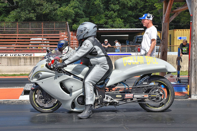 A motorcycle racer in a gray racing suit prepares to start a drag race, while another racer waits behind. A race official observes in the background.