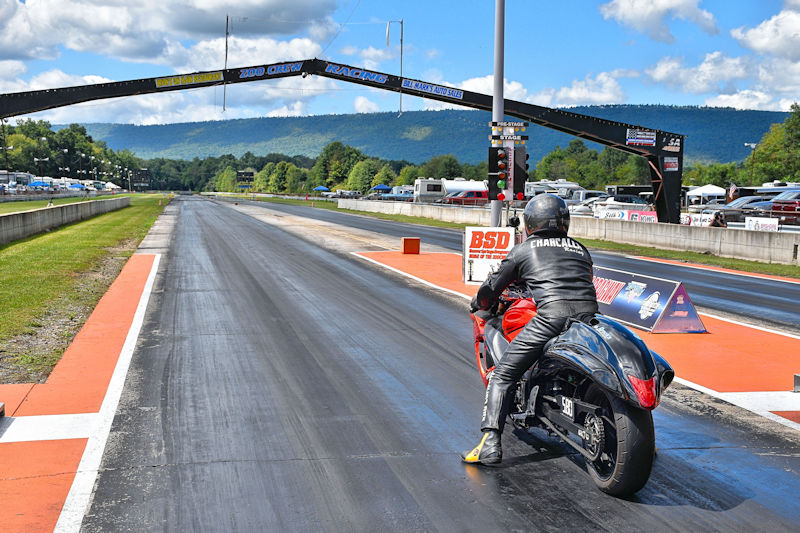 A motorcycle racer in leather gear prepares to launch from the starting line at a drag strip, with a clear view of the racing track and mountains in the background.