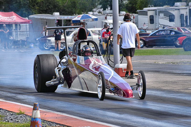 A drag racing car with a sleek design and large rear tire, accelerating down a racetrack, surrounded by smoke. A crew member stands nearby, observing the race preparations.