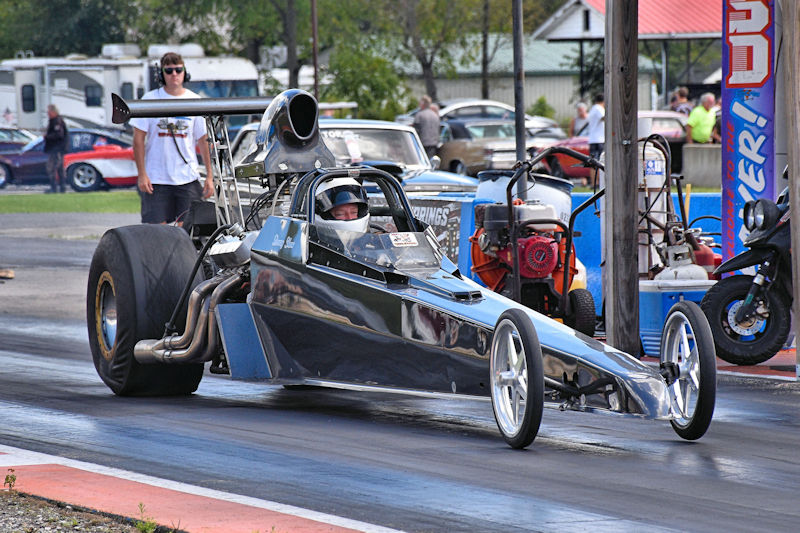 A shiny drag racing car accelerating down a track with a driver wearing a helmet, while spectators watch in the background.