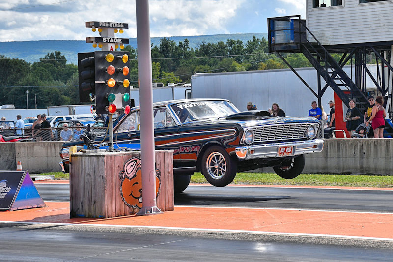 A classic car performing a wheelie at a drag racing event, with spectators in the background and a starting line with traffic lights visible.