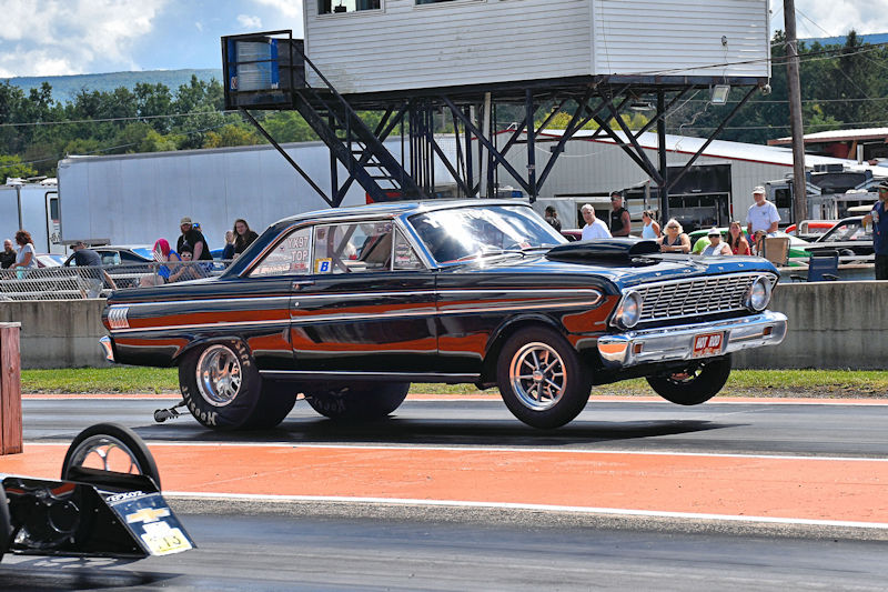 A classic black muscle car performing a wheelie on a drag strip, with spectators watching from the sidelines.
