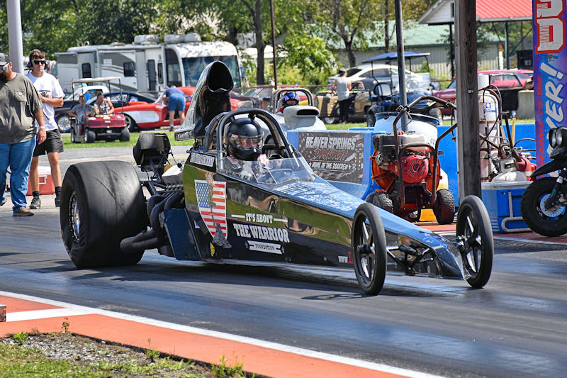 A drag racing car labeled 'THE WARRIOR' with an American flag, speeding down a racetrack, with spectators and additional vehicles in the background.