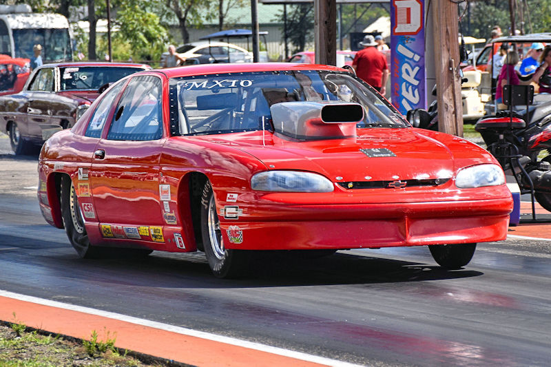 A red drag racing car with a large hood scoop, speeding down a racetrack with spectators in the background.