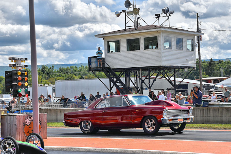 A vintage red car racing on a drag strip in front of a timing tower and spectators under a partly cloudy sky.