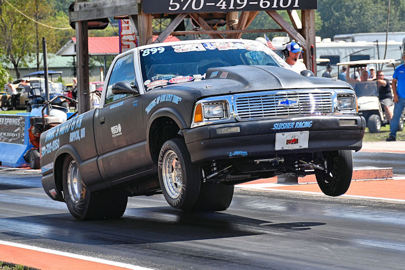 A black Chevrolet pickup truck performing a wheelie on a drag strip during a race event.