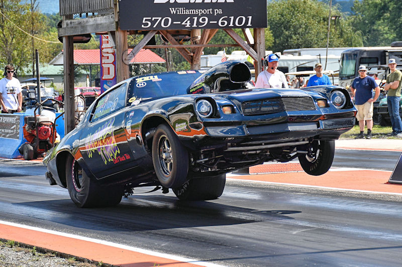 A black muscle car performing a wheelie on a drag strip, with spectators in the background watching the race.