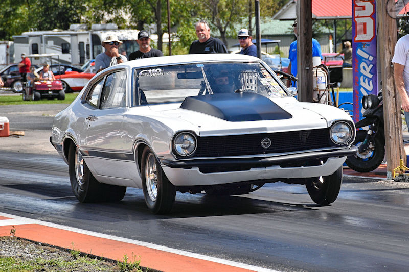 A white drag racing car with a black hood racing down a track, with spectators in the background.