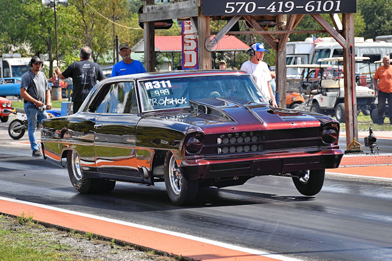 A black and red classic race car on a drag strip, with spectators in the background and a start signal structure overhead.