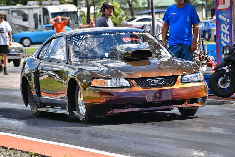 A modified racing car with a distinctive paint job, featuring a prominent hood scoop, is on a drag racing track. In the background, spectators are gathered while additional vehicles are parked.