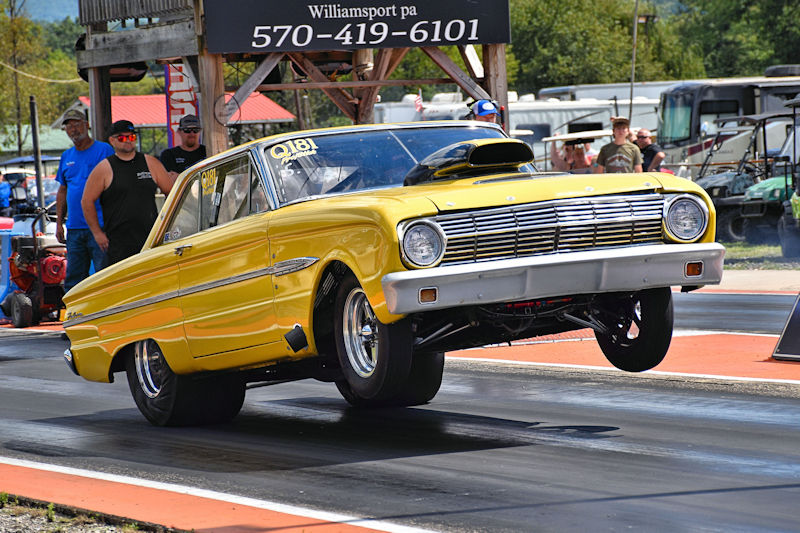 A bright yellow classic car performing a wheelie on a drag racing track, with spectators in the background.