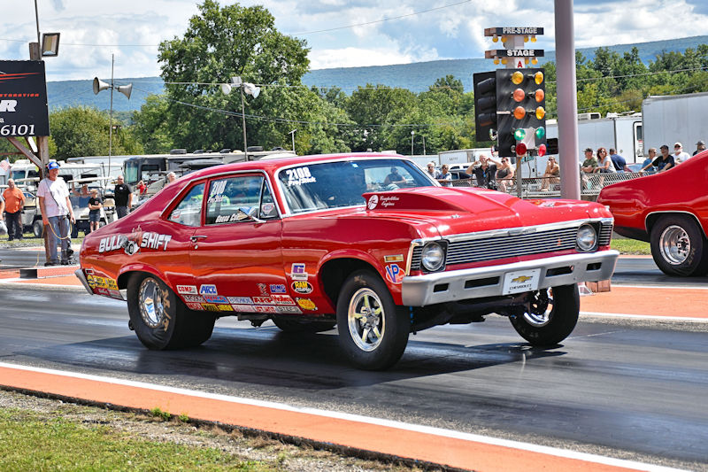 A classic red drag racing car on a track, with spectators in the background and drag racing lights in the foreground.
