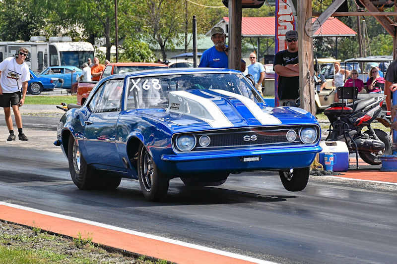 A classic blue Chevrolet Camaro SS with white stripes is performing a drag race on a race track. Spectators are visible in the background, along with parked cars and a canopy.