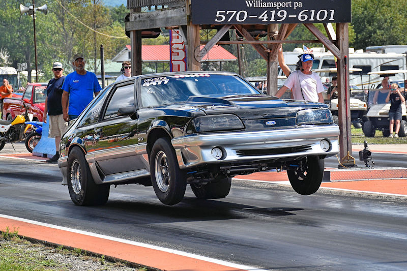 A black Ford Mustang performing a wheelie on a drag strip while spectators watch in the background.