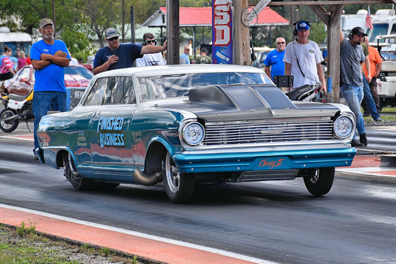 A vintage blue and silver drag racing car with a powerful hood, speeding down a racetrack, with spectators observing in the background.