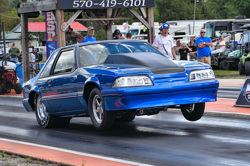 A blue drag racing car performing a wheelie on a racetrack, with spectators in the background.