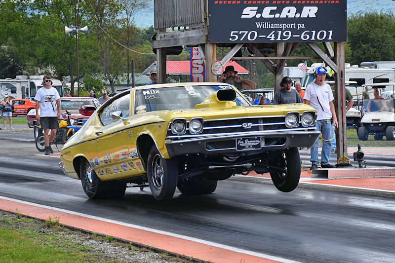 A yellow vintage muscle car performing a wheelie during a drag race on a race track, with spectators in the background.