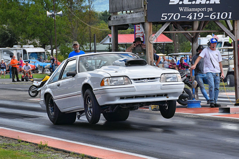 A white drag racing car performing a wheelie on a race track, with spectators in the background.