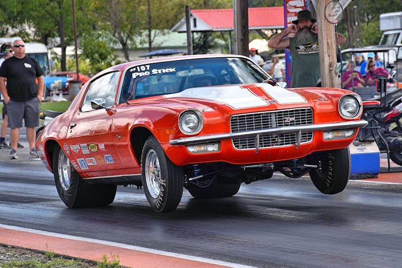 A classic orange muscle car performing a wheelie on a dragstrip, with spectators in the background.