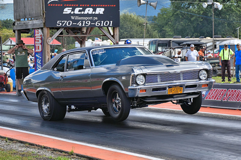A classic gray muscle car performing a wheelie on a drag strip, with spectators in the background and a sign for a local business visible.