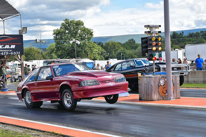 A red car performing a wheelie on a drag strip, with another car visible in the background and spectators watching nearby.