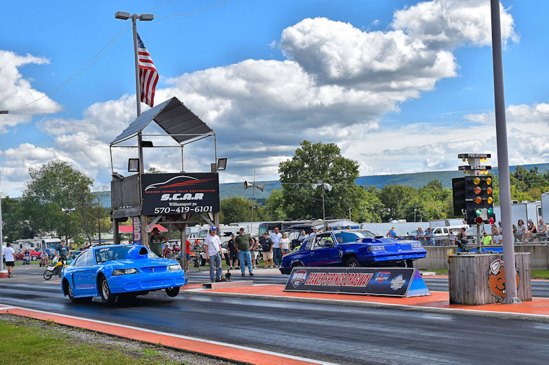 Two blue race cars accelerate down a drag strip during a race day, with spectators and a timing tower in the background.
