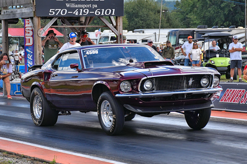 A classic 1969 Ford Mustang racing down a drag strip, showcasing its sleek design and powerful stance. Spectators watch from the sidelines as the car accelerates.
