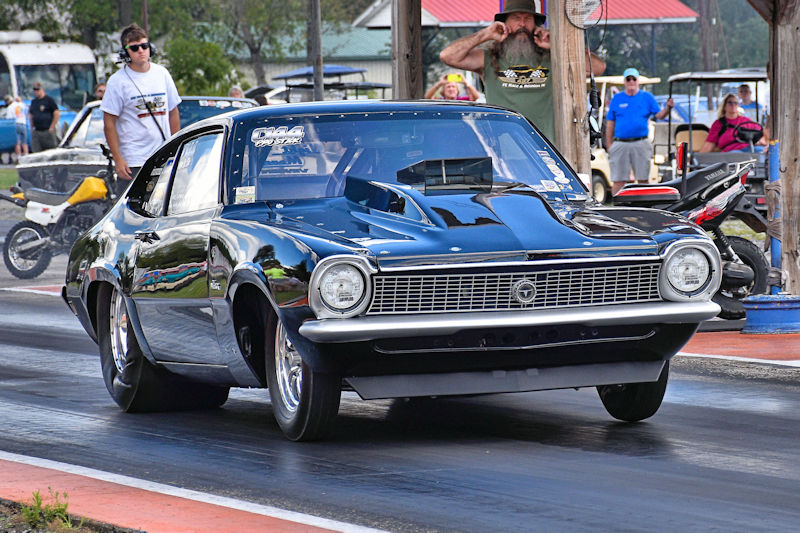 A classic black race car prepares to launch down a drag strip, with spectators in the background.