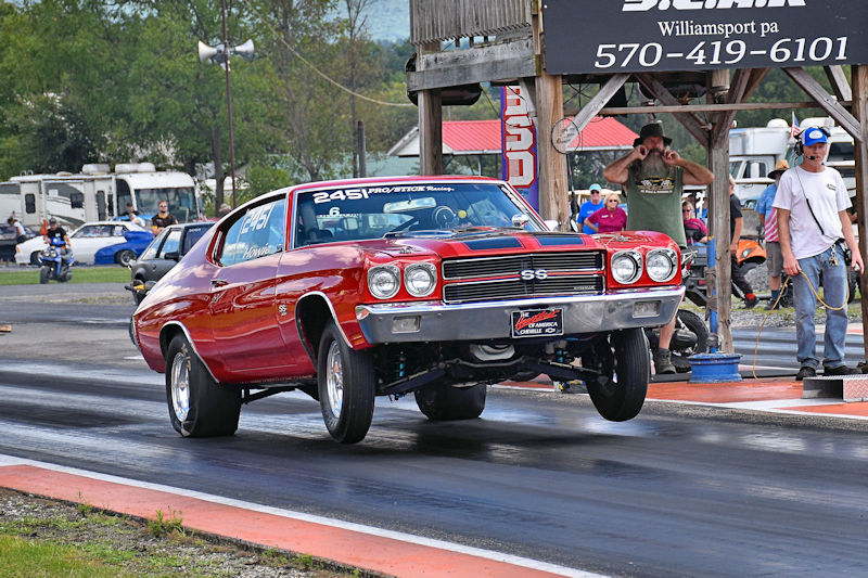 A classic red Chevrolet Chevelle SS performing a wheelie on a drag racing track, with spectators and trailers visible in the background.