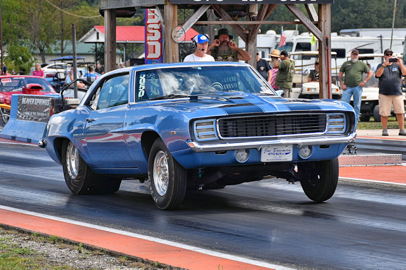 A classic blue muscle car, a 1969 Chevrolet Camaro, performing a wheelie on a drag strip during a race event. Spectators can be seen in the background, some watching the race and others in parked vehicles.