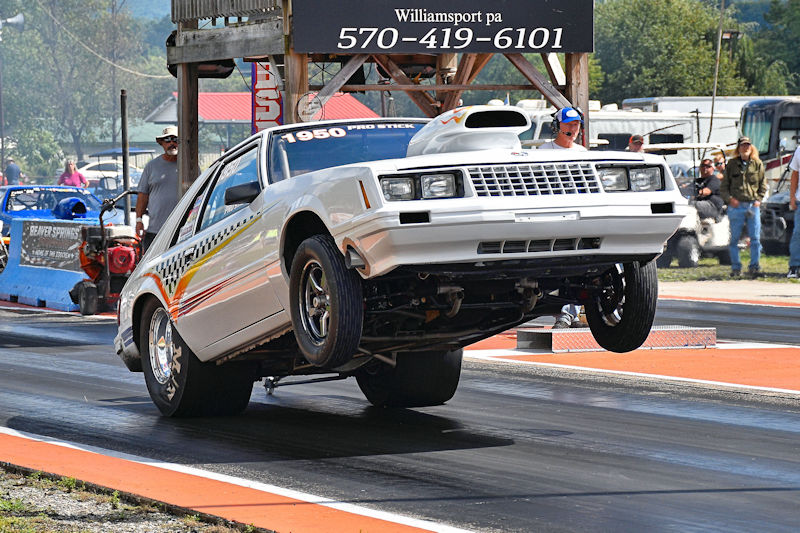A white race car performing a wheelie on a drag strip, with spectators in the background.
