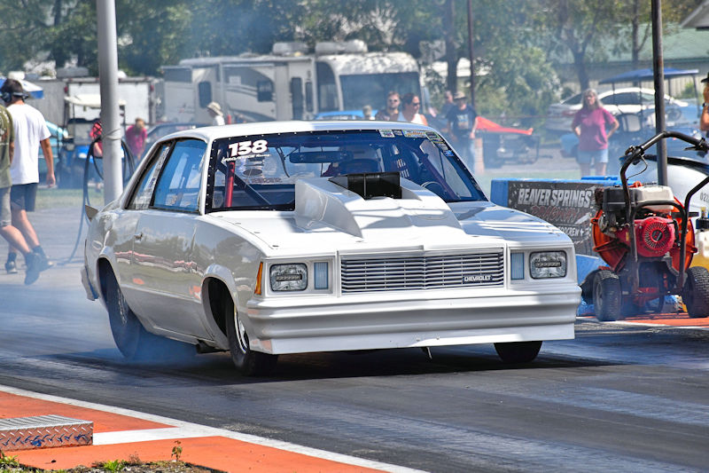 A white modified car with a large hood scoop racing down a drag strip, creating smoke from the tires, with spectators in the background.