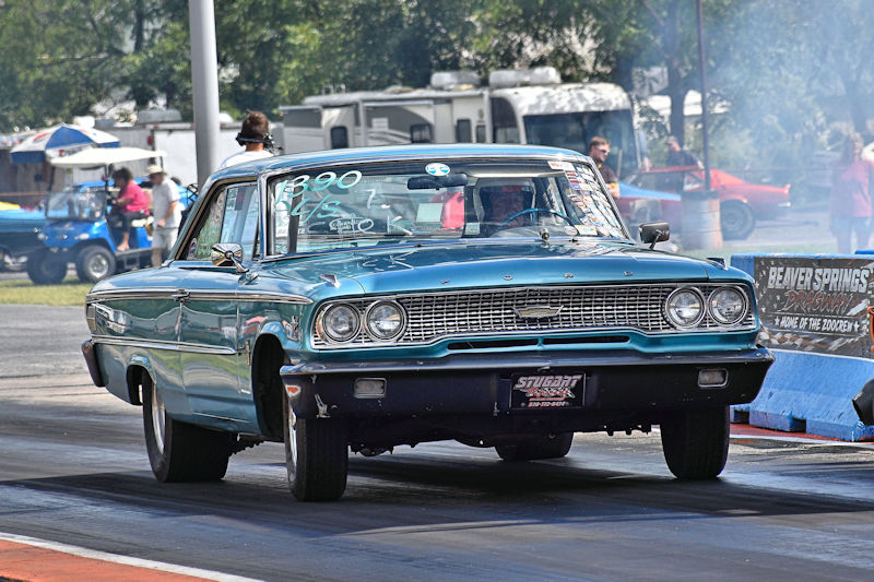 A classic blue Ford car racing down a drag strip with smoke trailing from the tires, surrounded by spectators and vehicles in the background.