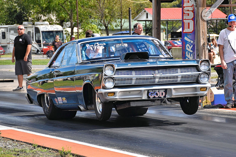A classic Ford car performing a wheelie on a drag racing track, with spectators in the background.