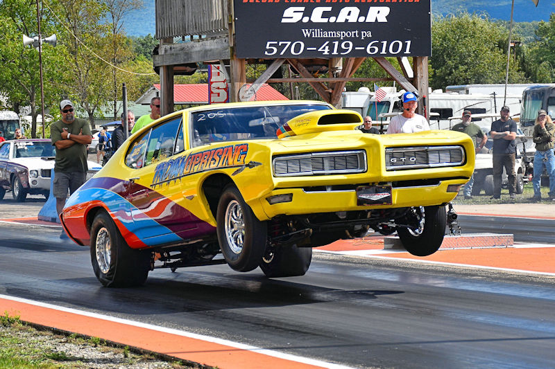 A yellow race car performing a wheelie on a drag strip, with spectators in the background and a sign for S.C.A.R. visible.