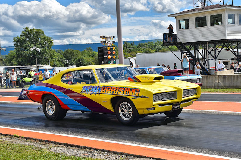 A colorful drag racing car with the text 'Indian Uprising' on its side, speeding down a drag strip with spectators in the background.