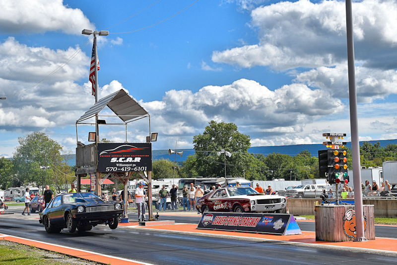 A drag racing scene featuring two cars on a race track with spectators in the background, under a blue sky with clouds. One car is launching down the track while the other is in a static position. There's a starting tower with an American flag and a scoreboard displaying race information.