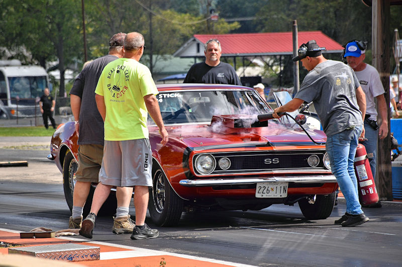 A group of individuals attending to a red vintage car at a drag racing event, with one person using a fire extinguisher near the car's hood, while others observe closely.