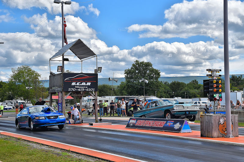 A blue race car is launching down a drag strip, with a classic green car in the background. Spectators are watching from the sidelines, and a race control tower is visible with a sign for the racing event.