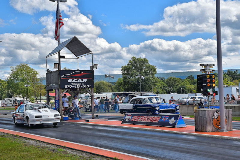 Drag racing scene featuring two cars at the starting line, with a race tower displaying a flag and timing lights in the background.