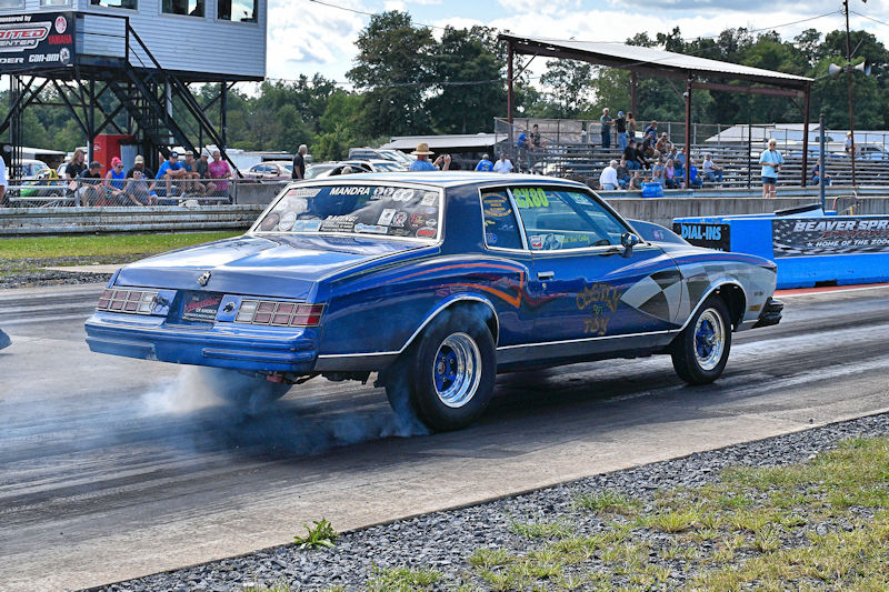 A blue racing car with a smoky burnout on a drag strip, with spectators in the background.