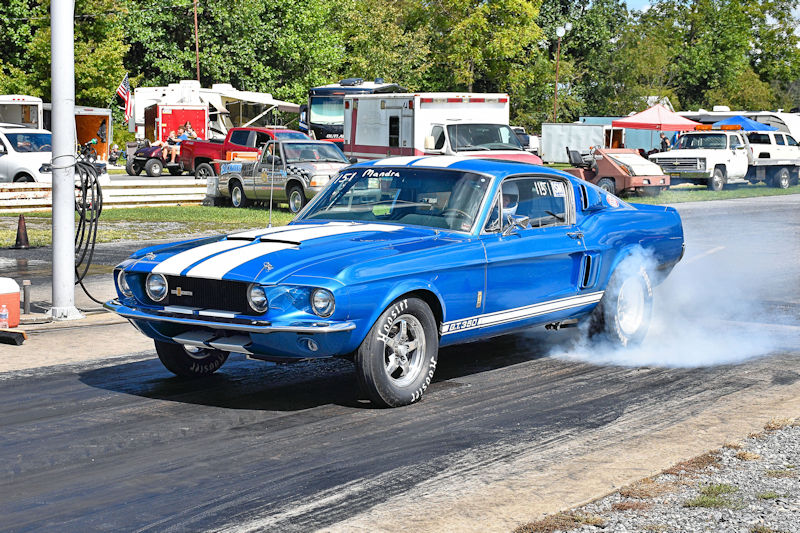 A blue 1967 Ford Mustang Shelby GT500 racing down a drag strip, leaving tire smoke behind.