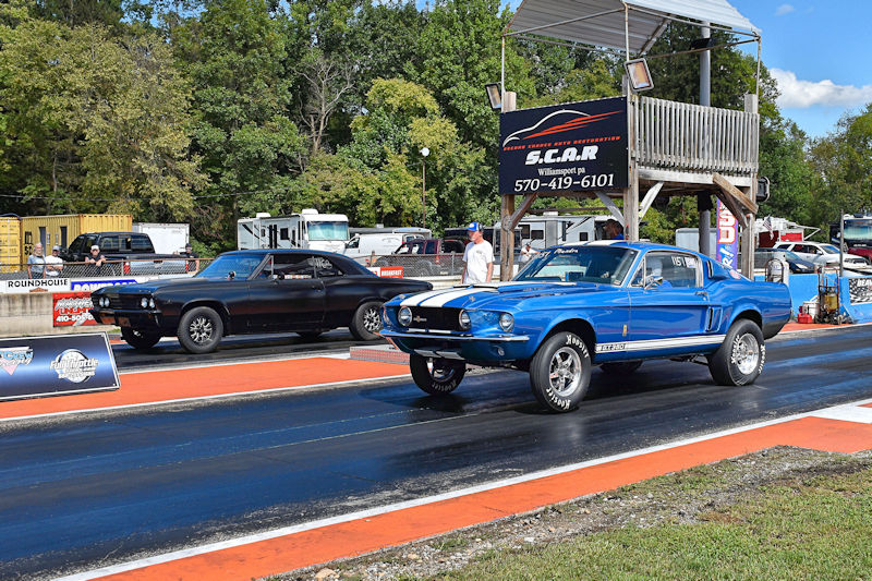 A blue classic muscle car racing on a drag strip alongside a black car, with a tree-lined background and a timing tower visible.