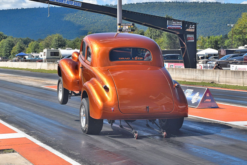 Vintage drag racing car with an orange finish launching down a drag strip in a scenic outdoor setting.