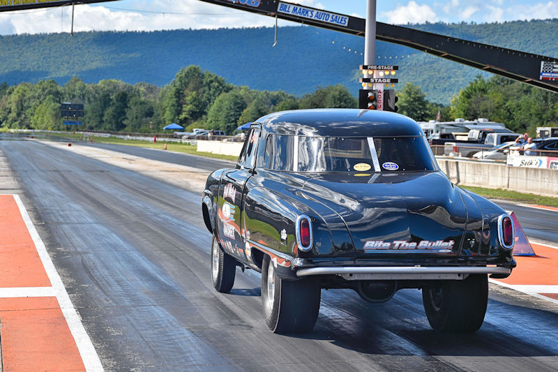 A classic black drag racing car launching down a drag strip with mountains in the background on a sunny day.