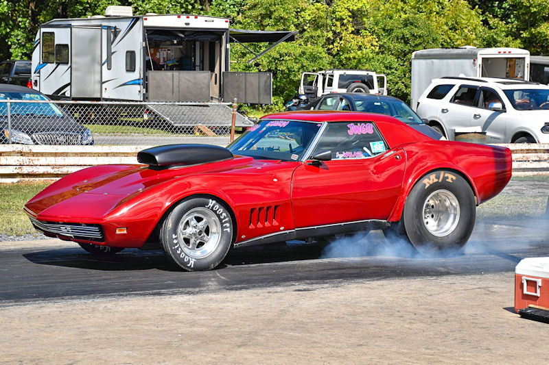 A red vintage racing car with a large hood scoop is performing a burnout on a drag strip, surrounded by parked vehicles and a trailer in the background.