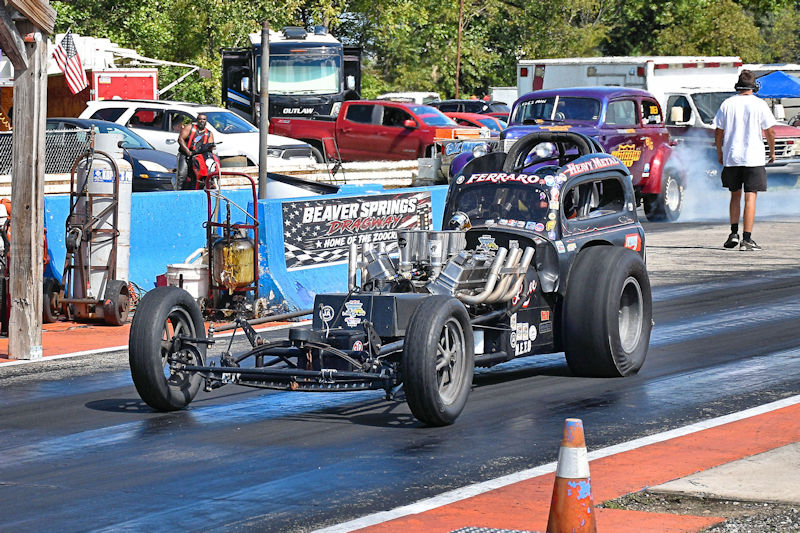 A vintage drag racing car on a racetrack, with smoke rising from the tires. The background features spectators, parked vehicles, and signage for Beaver Springs Dragway.
